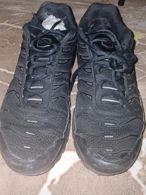 Nike Air Max Plus Tn helsvarta sneakers - Nike Air Max Plus Tn sneakers i helsvart med mesh- och syntetmaterial. Klassisk Tn-logga på hälen och vågformade detaljer på ovandelen. Skorna har snörning och en chunky sula som ger en sportig vibe.