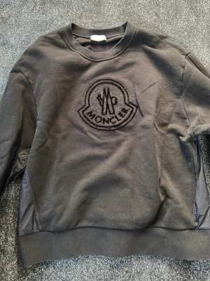 Moncler  - Svart sweatshirt från Moncler med stor broderad logga på bröstet. Tröjan har rund halsringning, ribbade muddar och är tillverkad i mjukt material. Perfekt för en stilren och avslappnad look.