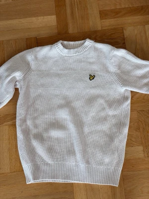 Vit stickad tröja från Lyle & Scott - Säljer en vit stickad tröja från Lyle & Scott med rund halsringning och det klassiska broderade logomärket på bröstet. Tröjan har ribbade muddar vid ärmslut och nederkant samt en snygg strukturstickning upptill.