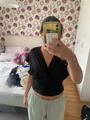 Svart topp - Svart omlottopp med djup v-ringning och bred midjeresår. Toppen har korta, lösa ärmar och knyt i nacken för en snygg passform. Perfekt till jeans eller kjol för en trendig look.