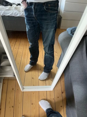 Blå jeans från Nudie Jeans - Säljer ett par klassiska blå jeans från Nudie Jeans med snygga slitningar och kontrastsömmar. Jeansen har fem fickor, raka ben och en cool läderpatch bak. Materialet är bomull och passformen är slim fit. Perfekt för dig som gillar en avslappnad och trendig stil.