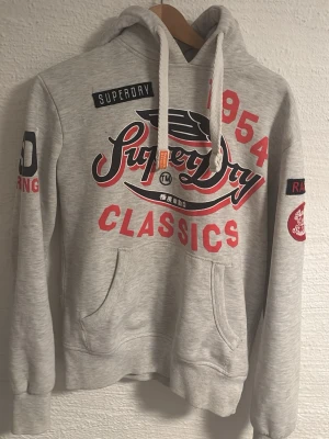 Superdry grå hoodie med tryck - Snygg grå hoodie från Superdry med stora röda och svarta tryck på bröstet och armarna. Klassisk känguruficka framtill, dragsko i huvan och broderade detaljer. Perfekt för en avslappnad streetstyle-look.