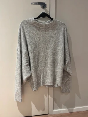 Ljusgrå oversized stickad tröja - Mysig ljusgrå stickad tröja med åtsittande midja. Tröjan har långa, lite bredare ärmar och ribbade muddar vid hals, ärmslut och nederkant. 