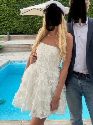 Studentklänning  - Supersöt vit axelbandslös kort klänning med volangig kjol och stora blommiga spetsdetaljer. Perfekt för student eller sommarfest. Klänningen har en figurnära överdel och fluffig kjol som ger en romantisk look.
