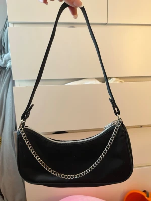 Svart axelväska med kedjedetalj - Trendig svart handväska i slät konstläder med silverfärgad kedja framtill och dragkedja upptill. Väskan har en smal axelrem och minimalistisk design som passar till många olika stilar. Perfekt accessoar för dig som gillar en clean look. På utsidan ser den ut som ny men på insidan är den lite smutsig. 