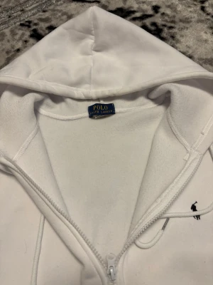 Vit hoodie från Polo Ralph Lauren - Snygg vit hoodie från Polo Ralph Lauren med dragkedja framtill, två fickor och klassisk luva med dragsko. Diskret broderad logga i svart på bröstet. Tillverkad i mjukt och bekvämt material, perfekt för en avslappnad stil.