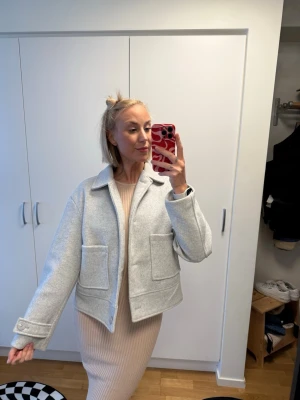 Ljusgrå jacka från 157 med fickor - Stilren ljusgrå jacka från 157 med klassisk krage och två stora fickor framtill. Jackan har en rak passform och är tillverkad i ett mjukt material som känns både modernt och bekvämt. Perfekt för lager-på-lager under kyligare dagar.