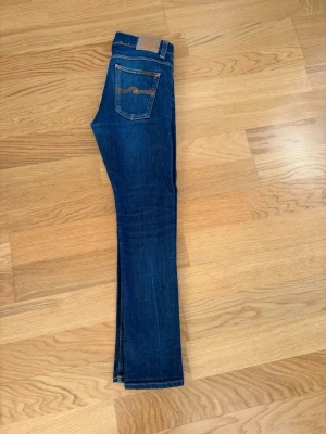 Mörkblå jeans från Nudie Jeans - Säljer ett par mörkblå jeans från Nudie Jeans med klassisk femficksdesign och kontrastsömmar. Jeansen har raka ben och Nudie-loggan broderad på bakfickorna. Tillverkade i denim med snygg tvätt och detaljer i läder på linningen.