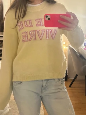 Gul stickad tröja med rosa tryck  - Söt gul stickad tröja med text i rosa bokstäver dekorerade med pärlor. Tröjan har rund halsringning, långa ärmar. Perfekt för dig som vill ha en mysig och trendig look.