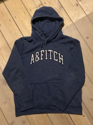 2000s Abercrombie & fitch hoodie - Mörkblå hoodie med stor huva och dragsko från Abercrombie & Fitch. Klassisk college-stil med broderad logga 'A&FITCH' i vitt på bröstet. Mjuk bomullsblandning och känguruficka framtill. Perfekt för en avslappnad och trendig look.