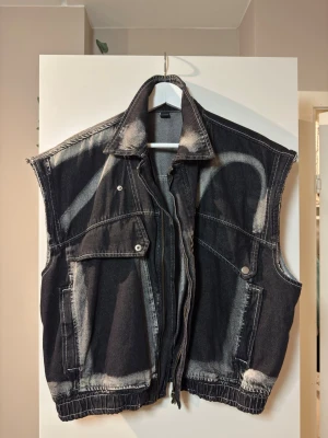 Oversized svart jeansväst med råa kanter - Säljer en oversized svart jeansväst med coola blekta detaljer och råa, avklippta ärmkanter. Västen har dragkedja framtill, två stora bröstfickor med lock och knappar samt två sidofickor. Materialet är kraftig denim och västen har en streetig vibe. (Kort och bred stil)