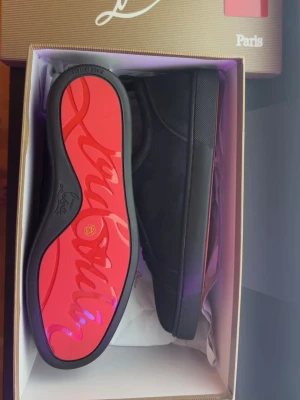 Svarta Christian Louboutin - Säljer ett par svarta låga sneakers från Christian Louboutin med klassisk röd sula och diskret logga undertill. Skorna har svart textil och mockaliknande material, svarta skosnören och metallöljetter på sidan. Perfekta för dig som gillar stilrena och exklusiva sneakers.