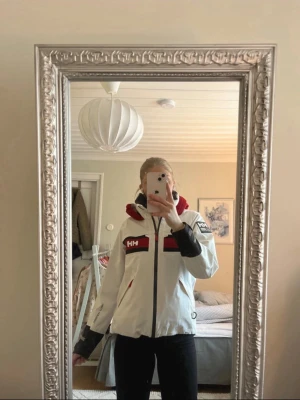 Vit och röd Helly Hansen vindjacka - Snygg vit vindjacka från Helly Hansen med röda och svarta detaljer. Jackan har dragkedja, huva med HH-logga, svarta muddar och flera fickor framtill. Perfekt för blåsiga dagar och har en sportig look med tydliga loggor.