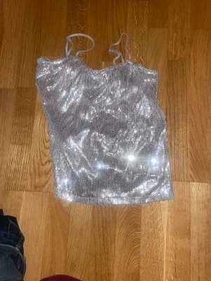 Glittrig silver linnetopp - Säljer en glittrig silverfärgad linnetopp med tunna axelband. Toppen har soprent snitt och är täckt av paljetter som reflekterar ljuset snyggt. Perfekt för dig som vill sticka ut och glänsa lite extra.