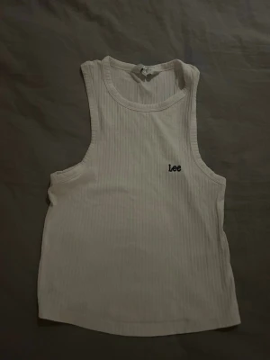 Vit ribbad linnetopp från Lee - Säljer en vit ribbad linnetopp från Lee med broderad logga på bröstet. Toppen är ärmlös och har en klassisk rund halsringning. Perfekt till sommaren och enkel att matcha med olika stilar.