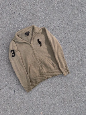 Grå stickad ziptröja Polo Ralph Lauren - Stickad beige ziptröja från Polo Ralph Lauren med stor svart polospelare broderad på bröstet och siffran 3 på ärmen. Tröjan har ribbade muddar, två fickor fram och hög krage. Perfekt för lager-på-lager och snygg till jeans. obs de finns en fläck 