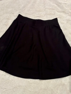 Svart kortkjol i mjukt tyg - Enkel och stilren svart kortkjol med utställd passform. Kjolen har bred midjeresår och är gjord i ett mjukt, stretchigt material som känns bekvämt. Perfekt att matcha med en crop top eller hoodie.