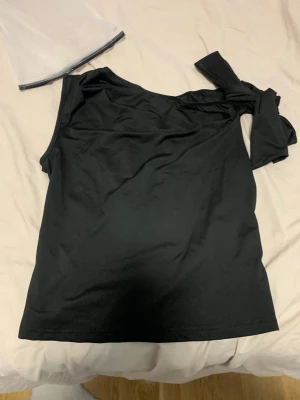 Svart oneshoulder topp med knyt - Snygg svart oneshoulder topp med knyt på axeln. Tillverkad i mjukt och stretchigt material som ger en tight passform. Perfekt för dig som vill ha en enkel men ändå trendig look.