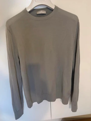 Ljusgrå stickad tröja i merinoull från Tiger of Sweden - En stilren ljusgrå stickad tröja från Tiger of Sweden med rund halsringning och långa ärmar. Tröjan har en klassisk passform och är tillverkad i ett tunt, mjukt material (merinoull) som passar perfekt till lager-på-lager. Enkel och tidlös design.