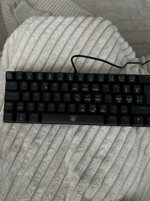 Svart mekaniskt tangentbord med sladd - Säljer ett svart mekaniskt tangentbord från Delux med kompakt design och nordisk layout. Tangentbordet har USB-kabel och tydliga vita tecken på knapparna. Perfekt för gaming eller skrivbordsarbete där du vill ha en smidig och stilren look.