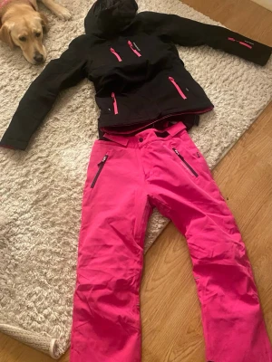 Rosa täckbyxor och jacka set från Swedemount - Snygga och funktionella rosa täckbyxor och jacka  från Swedemount med svarta dragkedjor och flera fickor. Perfekta för vinterns alla äventyr och håller dig varm och torr i backen. Byxorna har en modern passform och är tillverkade i slitstarkt material.