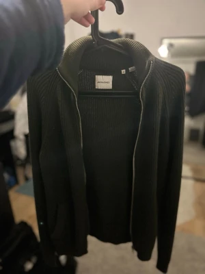 Olivgrön ziptröja från Jack & Jones - Snygg olivgrön ziptröja från Jack & Jones med ribbad struktur och hög krage. Tröjan har dragkedja framtill och två fickor. Perfekt för lager-på-lager och enkel att matcha med jeans eller chinos.