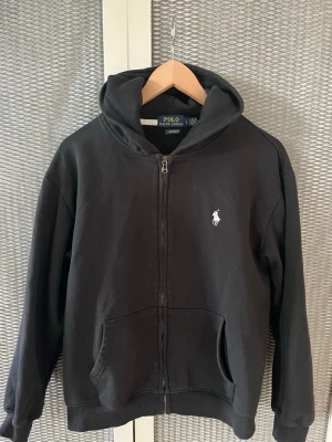 Svart hoodie/zip tröja från Ralph Lauren - Svart hoodie med dragkedja/zip up tröja från Ralph Lauren. Klassisk design med vit broderad logga på bröstet, huva med dragsko och känguruficka framtill. Mjuk bomullskänsla och ribbade muddar vid ärmslut och nederkant. Perfekt för en chill och stilren look. Storlek L fast sitter som S! SKICKA BUD