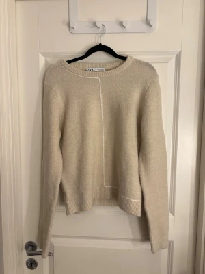Beige stickad tröja från ZARA - Säljer en beige stickad tröja från ZARA med rund halsringning och en tunn vit linje som detalj framtill. Tröjan har långa ärmar och ribbade muddar vid ärmslut och nederkant. Perfekt för dig som gillar enkel och stilren design.