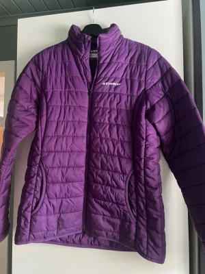 Lila pufferjacka från Everest - Snygg lila pufferjacka från Everest med hög krage och dragkedja framtill. Jackan har två fickor med dragkedja och quiltad design. Perfekt för kalla dagar och ger en sportig look. Materialet är lätt och isolerande. Liten i storlek. Har lagts på ena armen. 