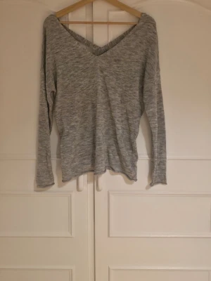 Grå långärmad tröja med V-ringning - Mysig gråmelerad långärmad tröja med djup V-ringning både fram och bak. Baktill finns snygga korsade band som ger en extra detalj.