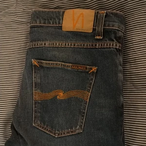 Blå jeans från Nudie Jeans - Säljer ett par klassiska blå jeans från Nudie Jeans med snygga slitningar och kontrastsömmar. Jeansen har fem fickor, orange broderi på bakfickan och patch med logga i midjan. Perfekta för en avslappnad och trendig look.