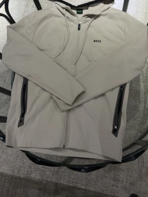 Beige hoodie med dragkedja från BOSS - Snygg beige hoodie från BOSS med dragkedja framtill och svarta detaljer vid fickorna. Hoodien har huva med dragsko och diskret BOSS-logga på bröstet. Tillverkad i mjukt material som känns skönt mot huden. Perfekt för en avslappnad och stilren look.