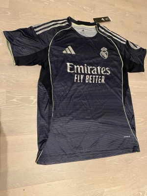 Real Madrid bortatröja Adidas - Snygg mörkblå Real Madrid bortatröja från Adidas med vita detaljer och klubbmärke på bröstet. Tröjan har rund hals, korta ärmar och tre klassiska Adidas-ränder på axlarna. Tillverkad i lätt och ventilerande material, perfekt för fotbollsträning eller match.