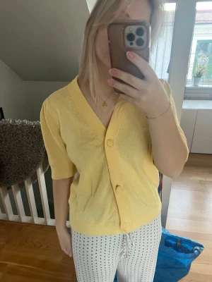 Gul kortärmad kofta med knappar - Söt gul kofta med korta ärmar och djup v-ringning. Koftan har bred ribbad kant och stora knappar framtill. Perfekt att slänga över en topp för en fräsch look. Materialet känns mjukt och lätt, passar bra till vår och sommar.