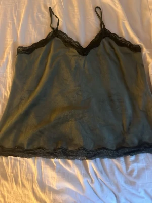 Grönt nattlinne med spetsdetaljer - Mörkgrönt nattlinne med tunna axelband och svarta spetsdetaljer längs urringning och nederkant. Tillverkat i ett glansigt, mjukt material som känns lyxigt mot huden. Perfekt för en avslappnad kväll hemma.