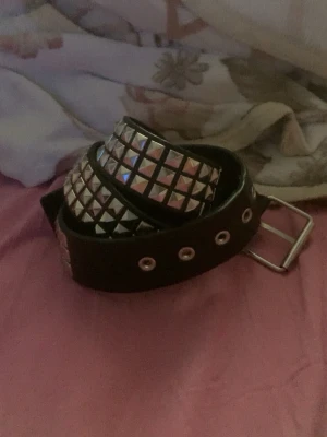Svart nitar bälte med silverdetaljer - Säljer ett svart bälte med silvriga fyrkantiga nitar och metallspänne. Bältet har flera hål för justering och ger en edgy vibe till outfiten. Perfekt accessoar för dig som vill sticka ut.