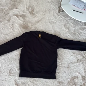 Svart hoodie från Emporio Armani - Snygg svart hoodie från Emporio Armani med klassisk passform och ribbade muddar. Perfekt för dig som gillar stilrena och exklusiva plagg. Diskret logga i nacken och mjukt material som känns skönt mot huden.