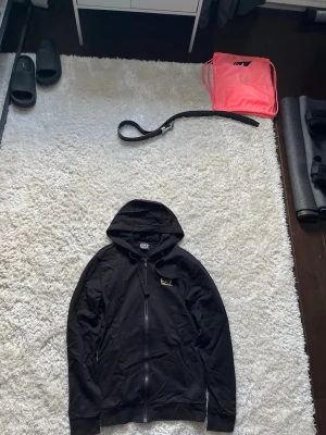 Svart hoodie från EA7 Emporio Armani - Snygg svart hoodie från EA7 Emporio Armani med dragkedja framtill och huva. Loggan i guld syns på bröstet och modellen har ribbade muddar vid ärmar och nederkant. Perfekt för en avslappnad och trendig look.