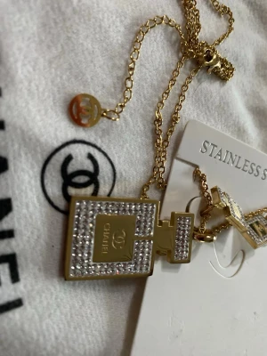 Guldfärgat Chanel halsband parfymflaska - Snyggt guldfärgat halsband + örhängen från Chanel med ett hänge i form av en parfymflaska, dekorerad med gnistrande stenar och logotyp. Kedjan har även en liten rund Chanel-logga. Tillverkat i rostfritt stål och perfekt för att lyfta din outfit.
