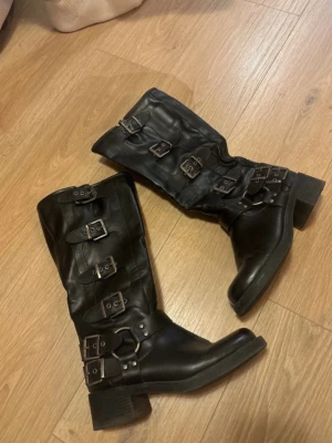 Svarta boots med spännen och klack - Säljer ett par svarta höga boots med chunky klack och flera coola metallspännen längs skaftet. Bootsens material är konstläder och de har en edgy look som passar till många olika stilar. Perfekta för dig som gillar detaljer och vill sticka ut.