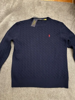Mörkblå stickad Polo Ralphlauren - Kabelstickad mörkblå tröja från Polo Ralph Lauren med rund halsringning och röd broderad logga på bröstet. Säljer pga att den är för liten. Den är knappt använd och pris kan diskuteras vid snabb affär! 