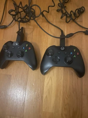 Xbox One med två svarta handkontroller - Säljer en Xbox One med två svarta handkontroller. Konsolen har en stilren, matt och räfflad design i plast och handkontrollerna har klassiska färgade knappar och USB-anslutning. Perfekt för gamingkvällar med vänner och familj.