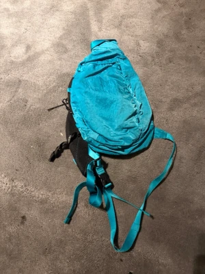 Turkos ryggsäck från C.P. Company - Säljer en turkos ryggsäck från C.P. Company med en unik rund svart detalj med loggan på sidan. Väskan har justerbara axelband och är tillverkad i ett lätt, slitstarkt material. Perfekt för dig som vill ha en cool och funktionell accessoar.