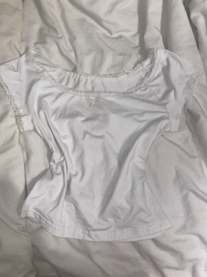 Vit topp med spetsdetaljer - Söt vit topp med korta ärmar och fina spetsdetaljer längs halsringningen och ärmsluten. Modellen är figurnära och har en feminin, enkel design som passar perfekt till jeans eller kjol.