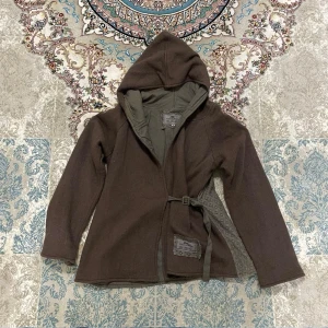 JDL kofta med huva – Dam - Mjuk damkofta från JDL med huva, omlottmodell och justerbart midjeband. Dekorativa spets-/meshpartier. Perfekt som lätt jacka eller kofta. Fint begagnat skick.  Storlek: M Mått (ca): 	•	Byst (armhåla–armhåla): ca 50 cm 	•	Längd: ca 60 cm 	•	Ärmlängd: ca 60 cm  Skick: Fint begagnat skick.