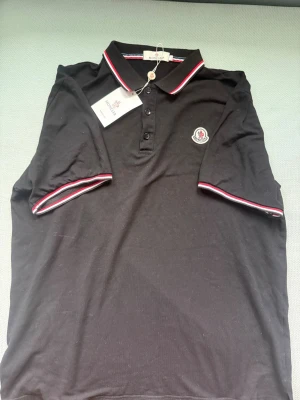 Svart Moncler pikétröja med röd/vit kant - Snygg svart pikétröja från Moncler med klassisk krage och knappar framtill. Tröjan har röda och vita detaljer på krage och ärmslut samt Moncler-logga på bröstet. Använd 3 gånger ungefär, väldigt bra skick. Skicka gärna frågor om tröjan