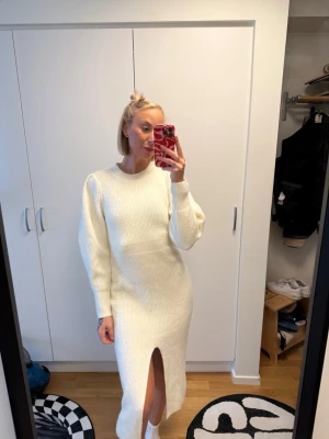 Vit stickad klänning med slits - Snygg vit stickad klänning med lång ärm och rund hals. Klänningen har en rak passform och en hög slits framtill som ger en cool touch. Perfekt för dig som gillar stilrena och trendiga plagg. Materialet ser ut att vara mjukt och ribbstickat.