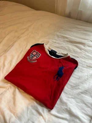 Röd t-shirt från Polo Ralph Lauren - Snygg röd t-shirt från Polo Ralph Lauren med blå logga broderad på bröstet och ett coolt klubbmärke på vänster sida. T-shirten har vita och mörkblå detaljer vid kragen och ärmarna. Perfekt för dig som gillar klassisk men sportig stil. 