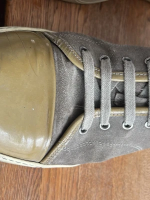 Bruna Lanvins - Sneakers i grå/brun mocka med beige/grön gummitå och matchande beige detaljer längs snörningen. Skon har grå skosnören och vit sula. Klassisk låg modell med metallöljetter och stilren design.