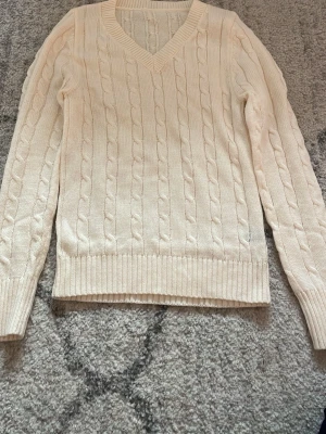 Vit kabelstickad tröja med V-ringning - Kabelstickad tröja i mjuk vit nyans med V-ringad hals och långa ärmar. Tröjan har ribbade muddar vid ärmslut och nederkant, vilket ger en klassisk och stilren look. Perfekt för lager på lager och enkel att matcha med olika outfits.